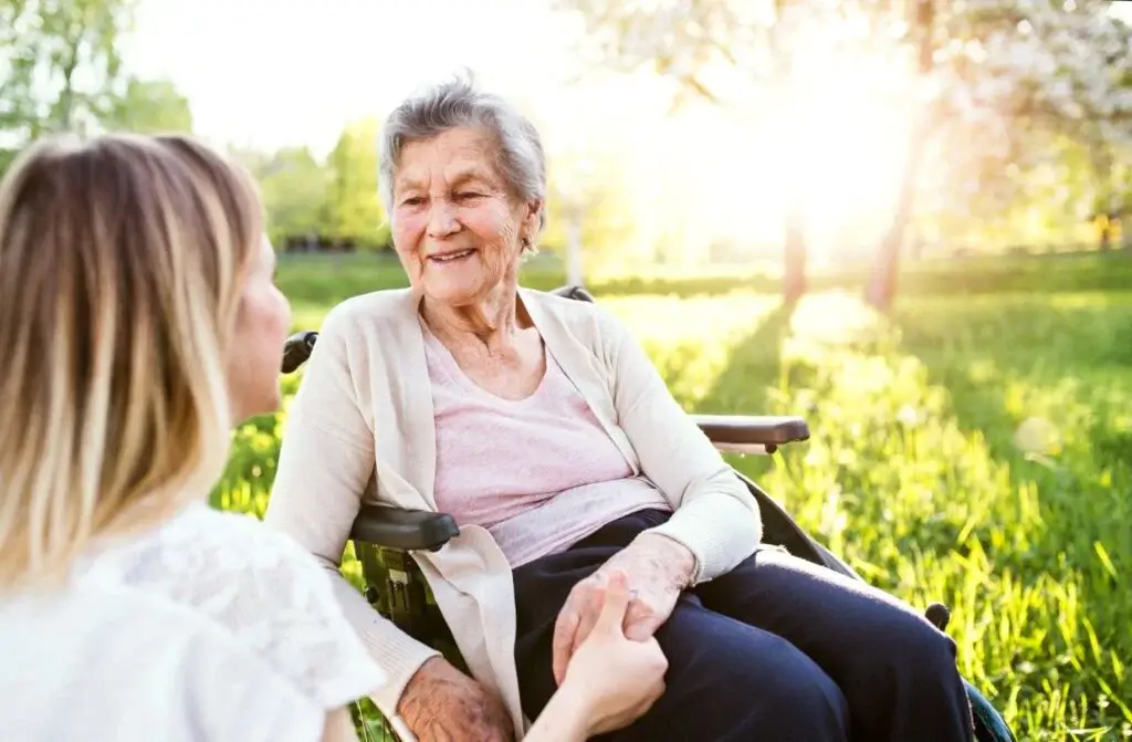 Woman kneeling next to an elderly woman in a wheelchair with grass and trees in the background.